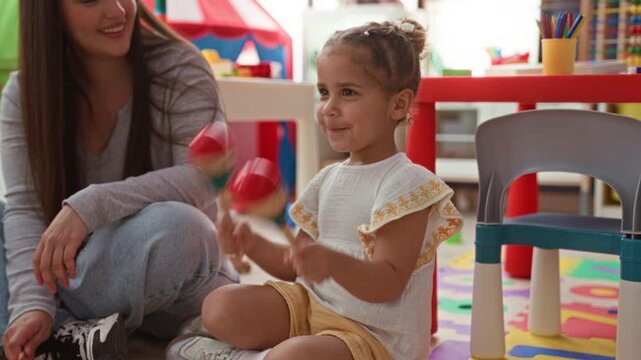 Girl shakes wooden maracas while sitting with teacher beside a red table and foam mat in a preschool building; joy playful learning.