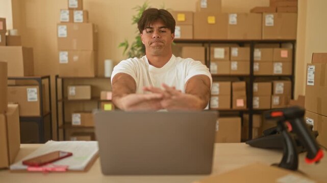 Man at laptop stretching hands forward among stacked cardboard boxes and shipping parcels inside a building; determination.