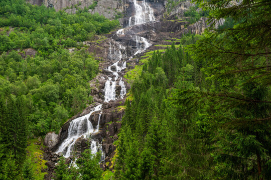 Dramatic Norwegian waterfall flowing through steep rock and lush green forest in summer.