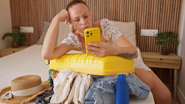 Woman overwhelmed by an overpacked yellow suitcase while check smartphone and prepare for an upcoming trip. Travel planning, digital lifestyle, decision fatigue ,modern vacation preparation concept.