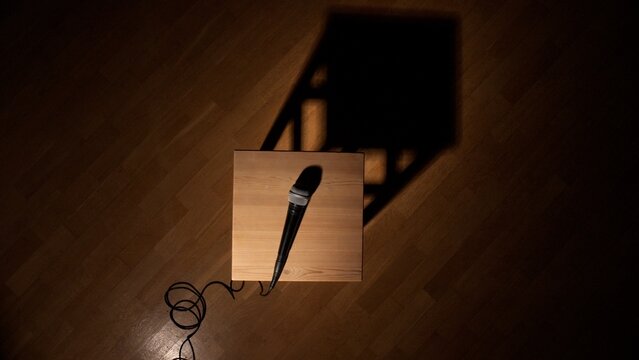 Microphone waiting on a wooden stool, casting a long shadow on the parquet floor, representing silence and anticipation before a speech or comedy act