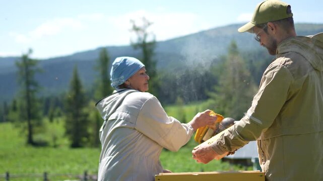 A cinematic medium shot of two beekeepers utilizing a traditional bee smoker to calm insects on a wooden honeycomb frame at a mountain apiary.