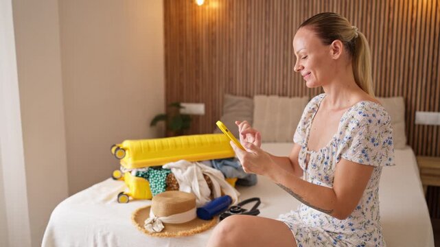Happy blonde woman sitting on her bed beside an open yellow suitcase filled with clothes, using her smartphone to plan an exciting summer vacation and book travel accommodation online.