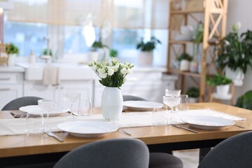 Fototapeta premium Stylish setting with vase of flowers, plates, cutlery and glasses on wooden table in kitchen