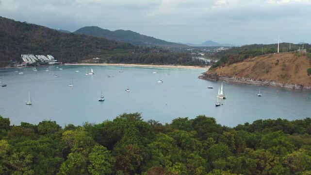 Aerial view of Nai Harn Beach capturing numerous sailboats anchored in the tranquil bay of Phuket, Thailand, surrounded by lush green hills and a sandy coastline. Drone flight zoom out panorama