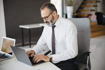 Obraz premium Experienced mature executive typing and analyzing data on a computer at his desk in a bright corporate workspace