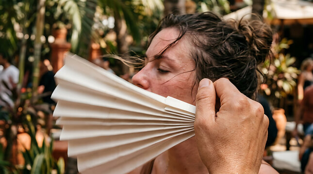 Woman fans herself to cool down during a hot summer day outdoors with tropical plants in the background