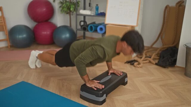Young hispanic man in olive top doing pushup on black aerobic step in a small home studio with exercise balls, battle ropes and a blue yoga mat; determination.