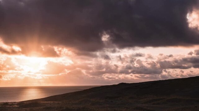 Dramatic golden sunbeams pierce through dark storm clouds over a tranquil ocean and silhouetted coastal landscape at dusk.