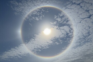 Obraz premium Sun halo with rainbow colors encircles bright sun in blue sky. Altocumulus clouds form pattern around atmospheric phenomenon. Light refraction creates optical illusion during daytime.
