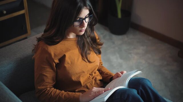 Young Hispanic woman reading a book on her home sofa, enjoying a peaceful moment of relaxation, learning, and personal development in a cozy indoor environment.
