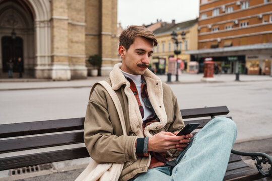 A stylish guy checks social media messages on a mobile phone while resting on a street bench outdoors in a historic European town center.