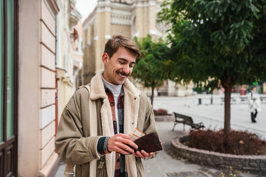 A cheerful tourist taking out paper money from a brown purse while standing outdoors in a European old town square.