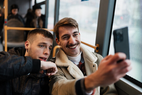 Happy male friends smiling and snapping photos on a mobile phone while riding urban public transportation during their daily commute.