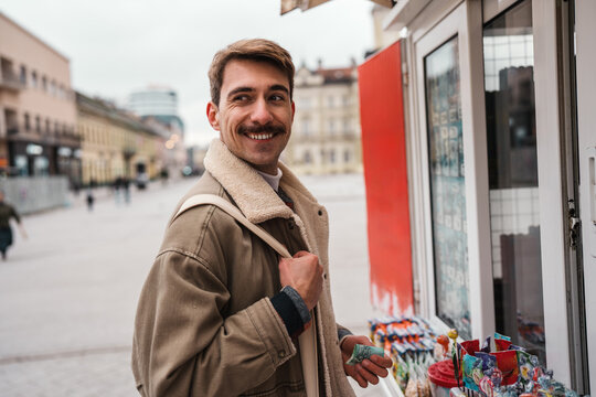 A smiling young guy dressed in a warm shearling jacket stands at an outdoor street booth to buy snacks with paper money on an urban square.