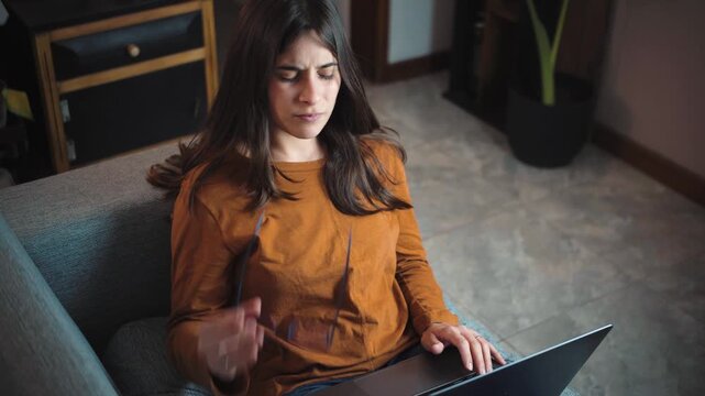 Young Hispanic woman working on her laptop from the sofa at home, then stopping due to severe neck pain caused by prolonged computer use and poor posture.
