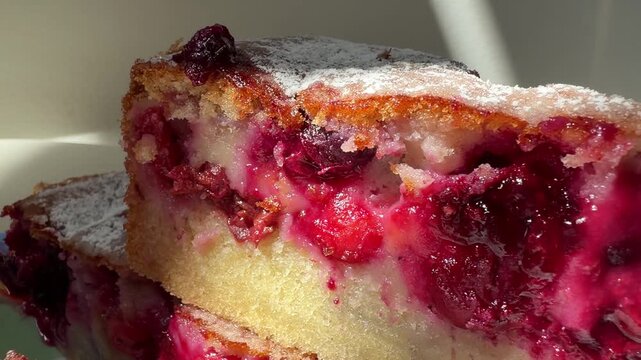 A homemade cherry sponge cake is shown whole, dusted with powdered sugar, and sliced to reveal its fruit-filled interior. The golden crumb and juicy cherries highlight a rustic dessert presented.