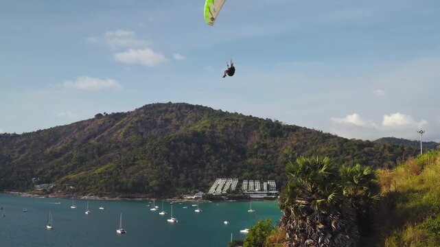 Phuket active sports and recreation. Paraglider soaring above beautiful coastal landscape of Nai Harn Beach, an aerial perspective of lush green mountains, tranquil ocean bay, and anchoring sailboats
