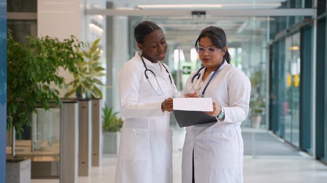 Two diverse female doctors interns discussing patient diagnosis in hospital hallway