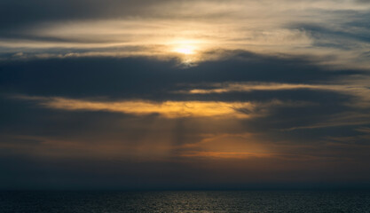 Fototapeta premium Golden sun rays breaking through heavy dark storm clouds over a dark open sea, creating a dramatic, atmospheric, and moody natural light spectacle over calm ocean waters at dusk