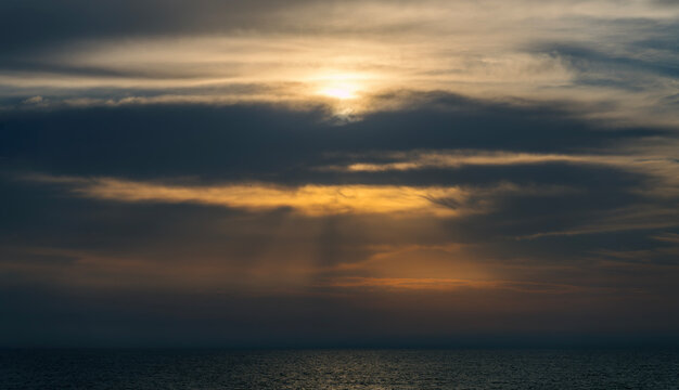 Golden sun rays breaking through heavy dark storm clouds over a dark open sea, creating a dramatic, atmospheric, and moody natural light spectacle over calm ocean waters at dusk