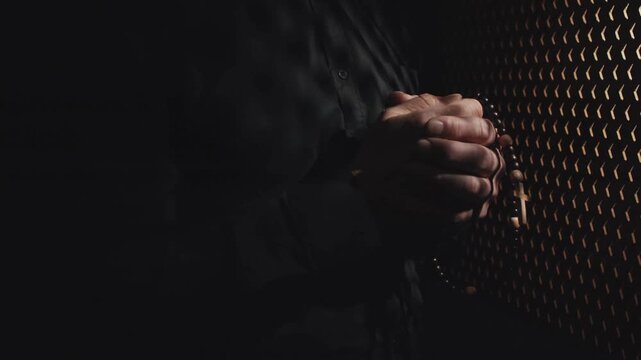Close-up of clasped hands in a dimly lit confessional booth, suggesting prayer or deep contemplation.