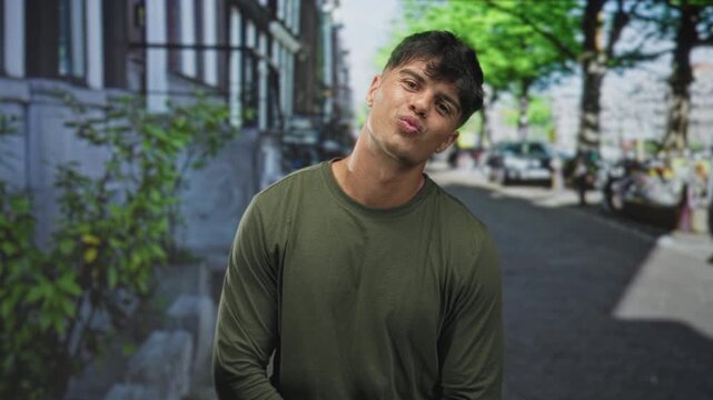 Young hispanic man in green longsleeve shirt puckers lips with tilted head on street; playful flirtation youthful charm.