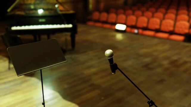 Closeup microphone and music stand on stage, soft spotlight hitting metallic grill, empty auditorium rows and grand piano out of focus suggest pre show calm
