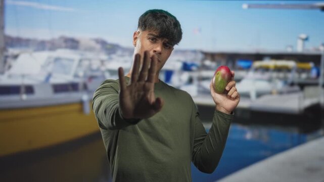 Man holding mango with palm out stop gesture at marina pier by docked boats, facing camera; caution restraint.