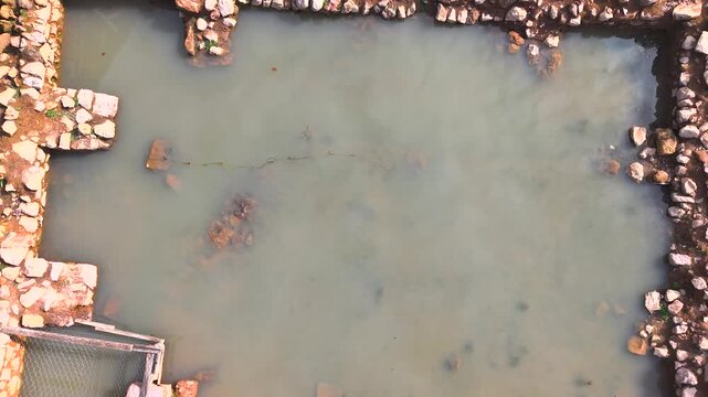 Smooth bird's-eye ascent begins with a close view of an ancient stone water reservoir filled with muddy water, gradually rising to reveal the surrounding archaeological landscape and historic architec