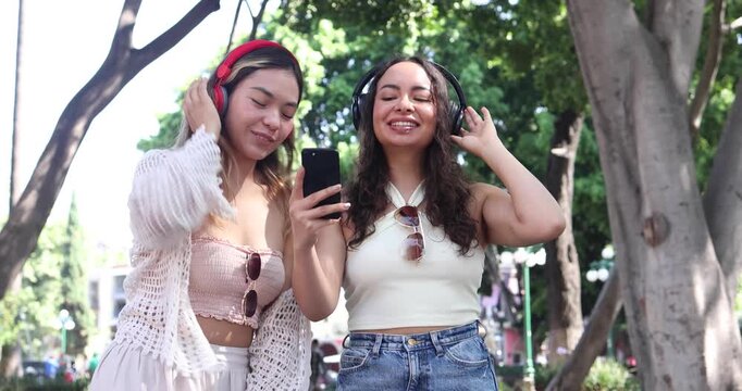 young latin couple of women friends using headphones and listening music in public park in Mexico Latin America