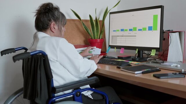 Determined disabled businesswoman in a wheelchair analyzing financial charts on her computer monitor, demonstrating inclusion and accessibility while working diligently from her home office desk