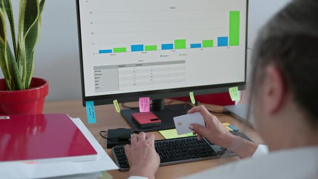 Unrecognizable businesswoman sitting at her office desk and entering credit card details on a computer keyboard to complete an online transaction while analyzing financial growth charts