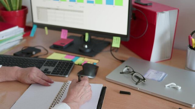 Senior businesswoman sitting at a messy office desk, taking notes in a spiral notebook while analyzing financial charts and data on a computer screen during a busy workday