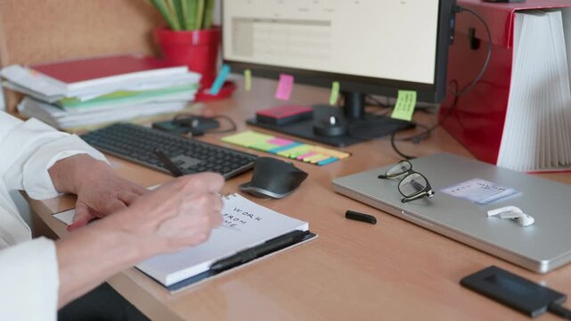 Business professional writing 'work from home' in a notebook while sitting at a cluttered desk with a computer and laptop, illustrating the concept of remote work and flexible scheduling