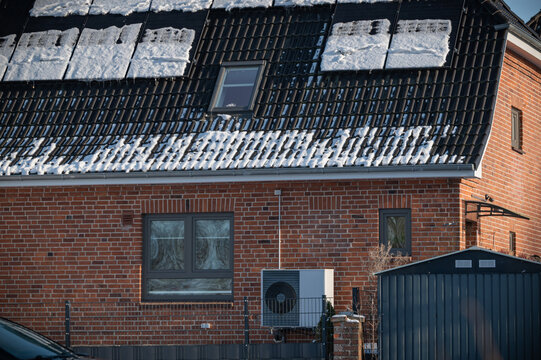 Modern brick house with solar panels in winter snow and outdoor heat pump unit for sustainable renewable energy.