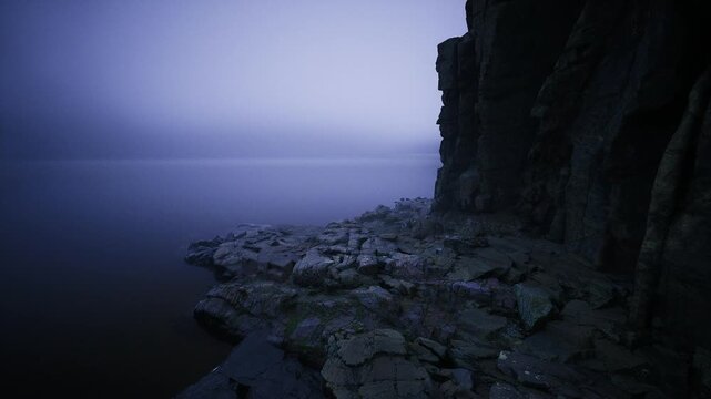 Jagged basalt promontory stretching into still sea, textured columns and rocky outcrops form stark foreground, placid water laps quietly while distant fog