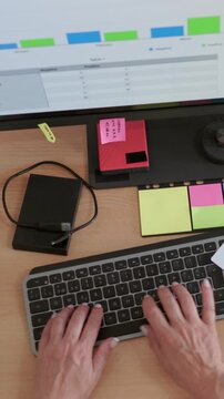 Overhead view of a financial analyst typing on a computer keyboard while examining charts and graphs on the screen, surrounded by sticky notes, a laptop, and other office supplies on a desk