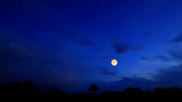 Full Moon on a twilight sky with landscape and tree silhouettes.
