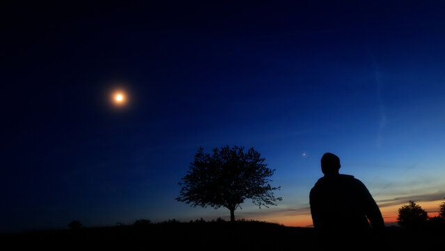 Silhouette of a person in nature observing night sky, Moon, stars and planets from a remote location.
