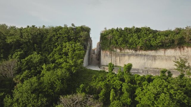 Zoom out aerial flight from narrow path carved through towering limestone cliffs, creating dramatic entrance to coastal landscape of Green Bowl Beach, Bali island, Indonesia. Remote travel destination