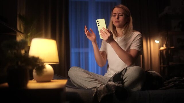 Beautiful young blonde woman looking at smartphone in bed at night with a shocked, open-mouthed expression and hand gesture of disbelief after reading breaking news