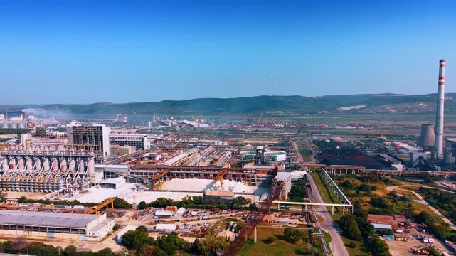 Territory of a large modern industrial plant with pipes producing smoke. Mountains in the backdrop.