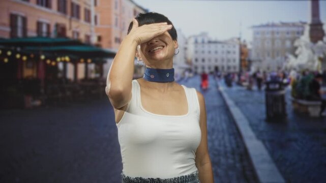Woman covers eyes with hand while smiling, wearing blue choker and white tanktop on a cobblestone street near a fountain and cafe; playful.