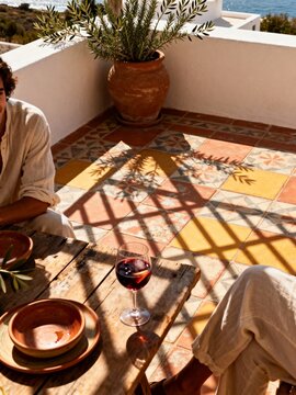 Mediterranean terrace with sea view and tiled floor. People enjoying drinks at a rustic wooden table on a sunny day. Summer vacation lifestyle concept