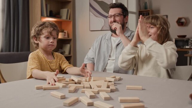 Caucasian family playing wooden block game at home, tower collapsing while kids and middle aged father react by laughing and clapping. Footage for family leisure and toy commercials
