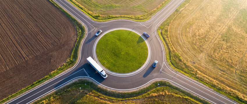 Aerial top view of a rural traffic roundabout with cars and a white truck navigating asphalt lanes surrounded by golden agricultural fields in warm sunlight for logistics and infrastructure