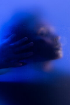 Artistic portrait of young black woman in blue light behind wet frosted glass. 