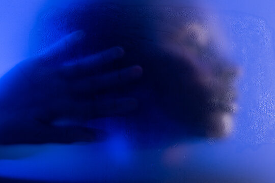 Emotional woman portrait behind wet frosted glass in blue light. Closeup of face and hand 