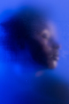 Anonymous beauty. Portrait of young black woman posing in studio with blue light behind wet steamy frost glass.